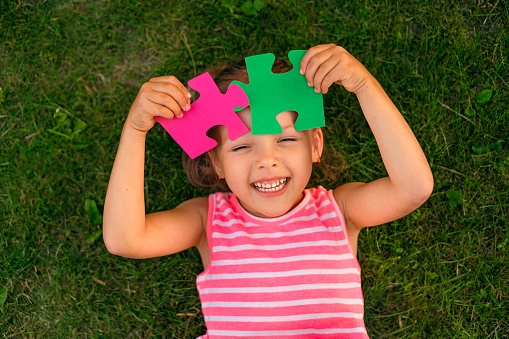 Funny laughing autistic girl playing with colored puzzle pieces lying on the grass in the garden
