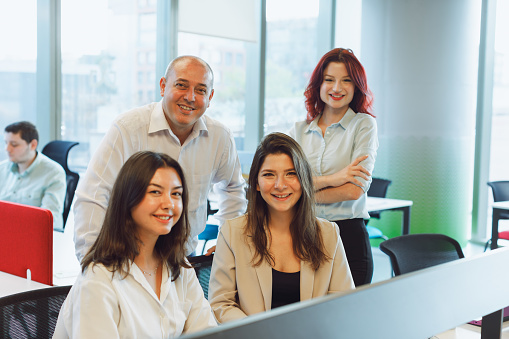 A vibrant portrait capturing the positive energy of a standing manager, CEO, and a group of two young businesswomen creative workers. They are all smiling and looking at the camera, exuding confidence and teamwork in an open modern office. In the background, their colleagues can be seen working diligently.