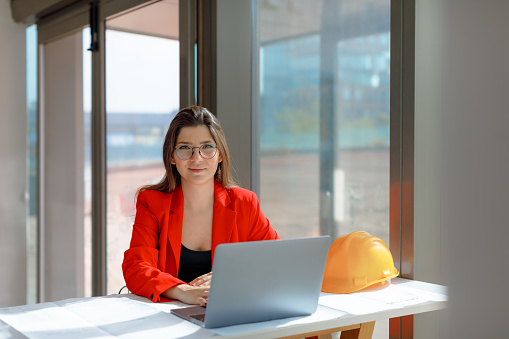 Portrait of the Happy and Successful beauty and satisfaction of a young and accomplished businesswoman. Dressed in a fashionable black shirt and a vibrant red blazer, she exudes confidence and professionalism. With her eyeglasses on, she smiles at the camera while working on her laptop and reviewing paperwork, surrounded by the creative atmosphere of a modern office. The sunlight streaming through the window adds a touch of brightness to the scene, reflecting her expertise in the fields of engineering, architecture, and business.