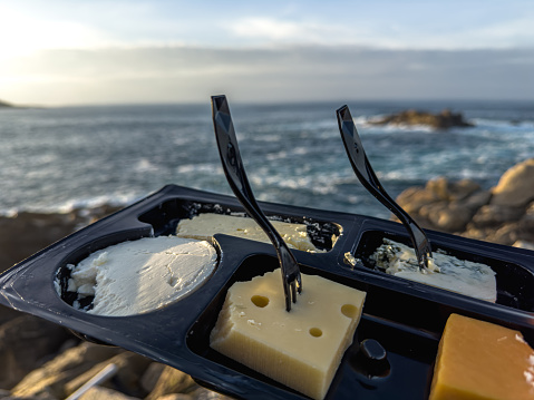 a close-up of a cheese selection presented on a sleek, black serving tray. Each piece of cheese is impaled with a modern black fork, ready to be enjoyed. Set against the backdrop of a serene seascape, the arrangement invites a moment of gastronomic pleasure amid the natural beauty of the ocean, with waves crashing against the rugged coastline under a soft sky.