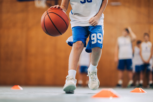 An unrecognizable young basketball kid is dribbling a ball on court during the training. Cropped picture of a young active boy dribbling a basketball on court during the training. Boy with basketball.