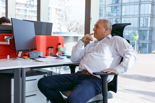 A portrait of a successful and experienced gray-haired mature businessman sitting at a desk in a modern open office. Engrossed in his work, he focuses on the computer monitor, analyzing statistics, commerce data, and marketing plans with precision and expertise.