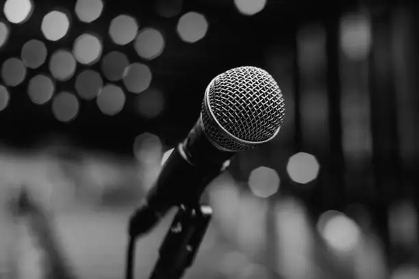 Black and white image of a microphone on an empty theater stage before a concert. Empty seats wait anxiously for the audience, while the stage lights remain blurred in the background. This microphone holds the promise of an emotional performance, where music and voices come to life. It's the critical piece of equipment that connects performers and audiences, delivering sound flawlessly at music events, conferences and comedy shows.