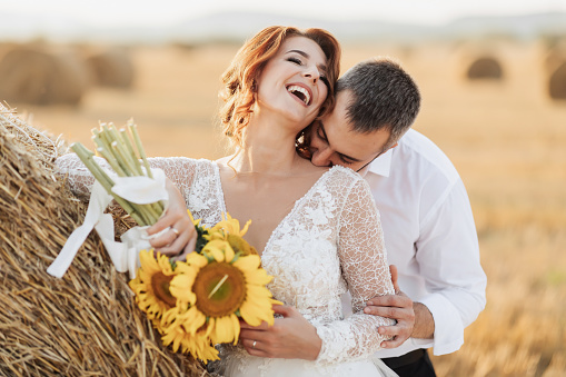 Wedding portrait of the bride and groom. The groom, tearing his shirt, stands behind the bride, near a bale of hay. Red-haired bride in a long dress with a bouquet of sunflowers. Stylish groom. Summer