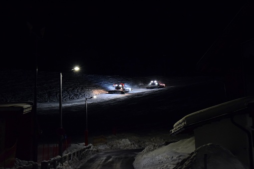 Two piste bashers working at compacting and smoothing snow in a ski resort at night.