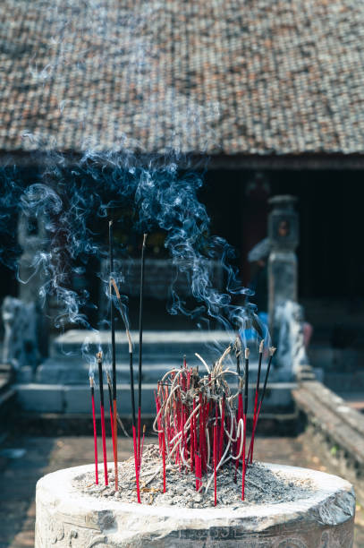 Asian woman is seen respectfully offering incense sticks in a traditional temple setting, with sun rays filtering through the smoke, creating a serene and spiritual atmosphere. The scene reflects a deep sense of spirituality and cultural tradition.