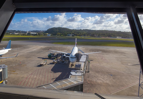 The image shows the view from the window of the Food Court at Recife/Guararapes International Airport - Gilberto Freyre, the main airport in the Northeast Region of Brazil, with a plane on board on the runway ready for boarding on a sunny day.