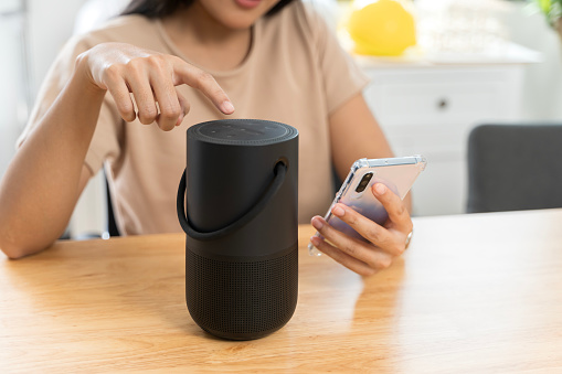 Close-up of a wireless speaker in front of a woman sitting on a work desk at home. Woman streams music or podcasts to smart speaker from mobile phone at home.