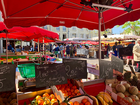 Horizontal closeup photo of fruit and vegetable stalls under red awnings and people shopping, walking through the market on a sunny day in Springtime. Saumur, Loire Valley, Maine-et-Loire, France, Europe. 20th April, 2019