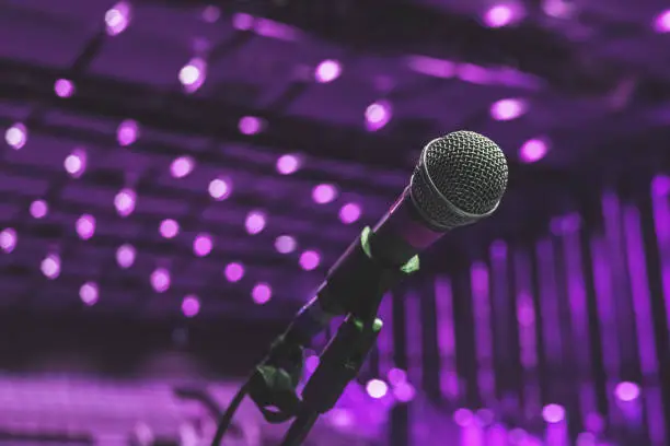 Microphone On The Theater Stage Before The Concert With Purple Blurred Lights. The stage is set, the lights are dimmed, and the anticipation fills the air as the microphone stands ready for its myriad of uses. From electrifying concerts to soulful music performances, captivating theater productions to engaging monologues, hilarious stand-up comedy to enlightening seminars and workshops, the microphone serves as the conduit for voices to be heard and stories to be shared. It amplifies the melodies of choirs and orchestras, accompanies the rhythmic beats of bands and solo artists, and empowers speakers to deliver impactful presentations and speeches. Whether it's a music festival, theatrical production, corporate event, or educational seminar, the microphone plays a vital role in shaping the auditory experience and connecting performers with their audience.