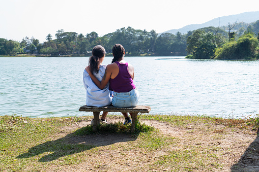 Back view of two diversities of ethnicity friends, young Asian female and African female, sitting on wooden chair enjoying their time in national park, looking into beautiful pond landscape, talking and spending relaxing time together in spring season.