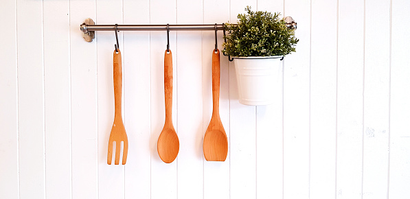 Brown wooden fork, spoon, turner or flipper and green plant in white stainless pot hanging and isolated on white wooden background with copy space. Kitchen utensil, Plant and Vintage object concept