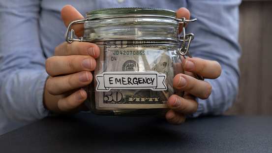 Unrecognizable woman holding Glass jar full of American currency dollars cash banknote with text EMERGENCY. Preparation saving money. Moderate consumption and economy. Collecting money