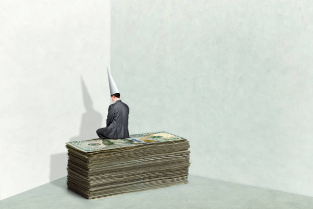 A man in a corner wearing a dunce cap sitting on top of a stack of money is guilty of financial illiteracy.