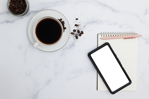 A cup of hot coffee sitting with a smart phone and a notepad with pen on a marble desk top complete this still life flat lay.