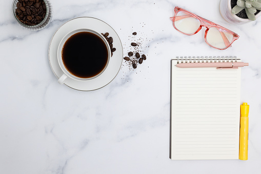 A hot cup of black coffee joins a busy desk occupied by a note pad, pen, glasses and highlighter.