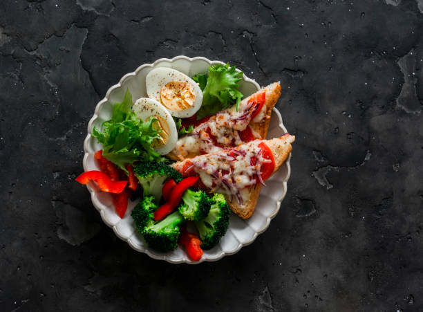 Delicious diet lunch, breakfast - boiled egg, fresh vegetable salad, broccoli and hot sandwiches with tomato and cheese on a dark background, top view