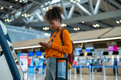 African woman holding passport using self Check-in kiosk machine getting airline ticket boarding pass at airport terminal. People travel on holiday vacation and global airplane transportation concept.