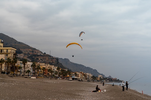 Letojanni, Italy - 29 January, 2023: fishermen on the beach at Letojanni with paragliders landing behind them after a flight from the Sicilian mountain tops
