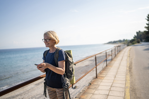 Mature woman texting on mobile phone while walking with backpack along the seashore on sunny day