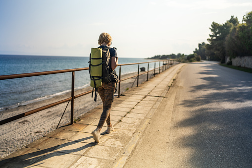 Rear view of mature woman hiking with backpack by the sea on sunny spring day