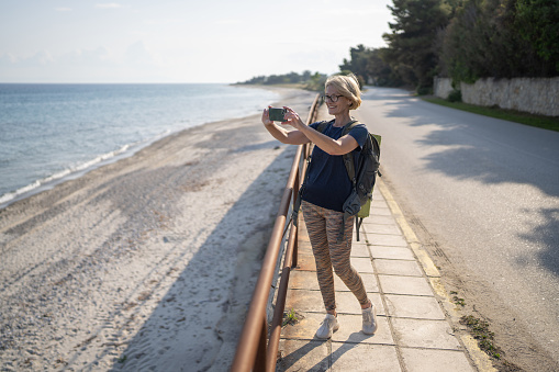 Full length shot of mature woman using mobile phone to take a photo of the sea on her hiking adventure
