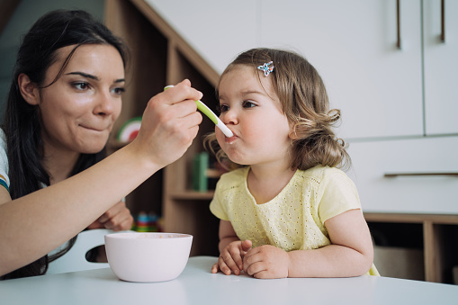 Shot of a happy young mother feeding her baby girl with a spoon at home.