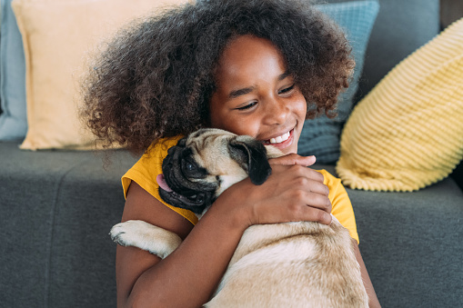 Shot of cute afro-american girl hugging her pug dog in the living room at home.