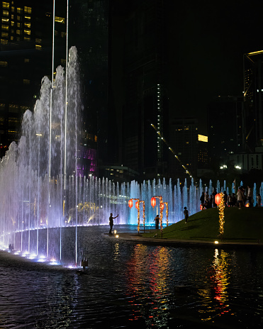 The pretty light and water fountain show takes place each night at the Petronas Towers . There are some unrecognizable tourists in silhouette in the image. Room for copy