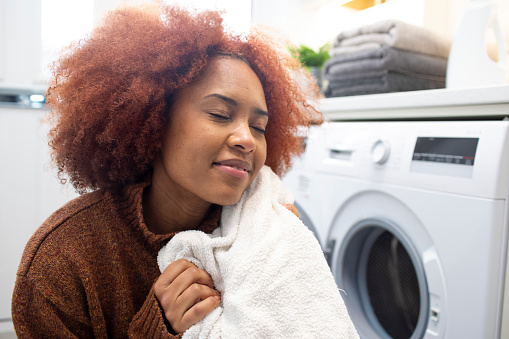 Young woman doing the laundry in a utility room at home. She is holding a clean toweling bathrobe close to her face.