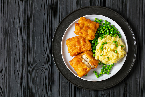 crispy fried fish fillet with mashed potatoes and green peas on plate on black wooden table, flat lay, free space