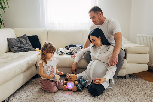 Parents are playing with their daughter in the living room. A pregnant woman is sitting on the floor next to a little girl and they are playing with stuffed toys