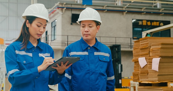 Young Asian woman worker and male manager walking checks stock inventory with tablet and discuss talk together in manufacturing factory. Working in manufacturing plant or production plant concept.