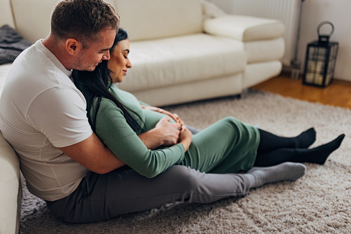 Husband and wife are sitting in each other's arms on the carpet in the living room. A man hugs his wife's pregnant belly