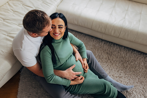 A man and a smiling, pregnant woman are sitting on the carpet in the living room. A man hugs his wife's pregnant belly