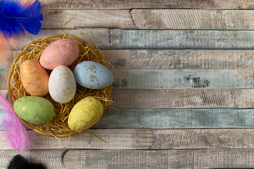 Flat lay composition of colourful painted easter eggs in a hay basket on wooden table. Happy Easter celebration