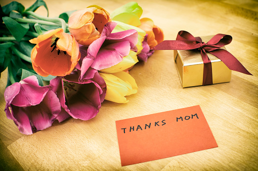 High angle view of golden gift box with tied bow on wooden background, close to the box is a greeting card. On left top corner is a bunch of tulips.