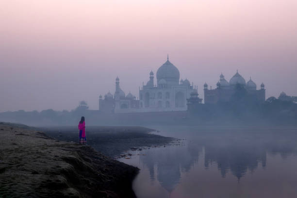 Taj Mahal,Agra,India-December 10 2023 : Taj Mahal in the beautiful morning sunrise and a woman is wearing Indian traditional clothes and standing by the river, Taj Mahal is the jewel of Muslims art in India and one of the universally admired masterpieces of the world's Heritage.