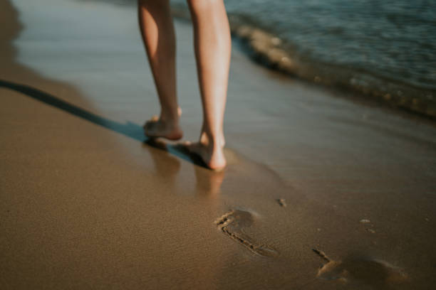 Woman walking along the shore leaving her footprints in the sand. The footprint is focused. Sunset at the beach. Take a walk to disconnect. Soft and pleasant waves. Beautiful woman legs. Relaxation in summer.
