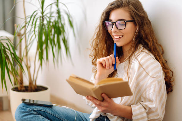 istock Young woman sits at home, using smartphone, learning, reading book. 2009816983