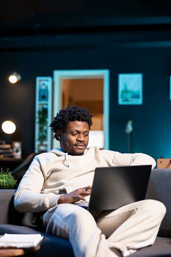 Smiling self employed man checking his emails on laptop while laying on couch. Cheerful remote employee working from home in bright stylish cozy apartment personal office, reading news on notebook