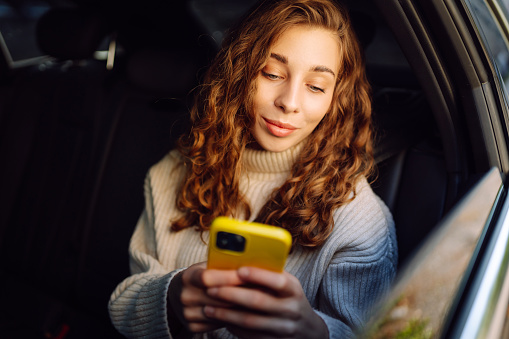 Beautiful young woman uses a smartphone while sitting in the back seat of a car. Woman talking on phone while traveling in the car. Internet and social media.