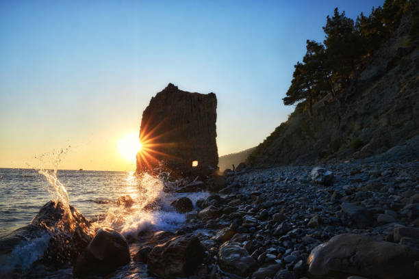 Sun dips below horizon, rocky coastline, nature beauty in serene tranquility. Waves gently lap against shore, while silhouetted cliffs stand majestically against dusky sky