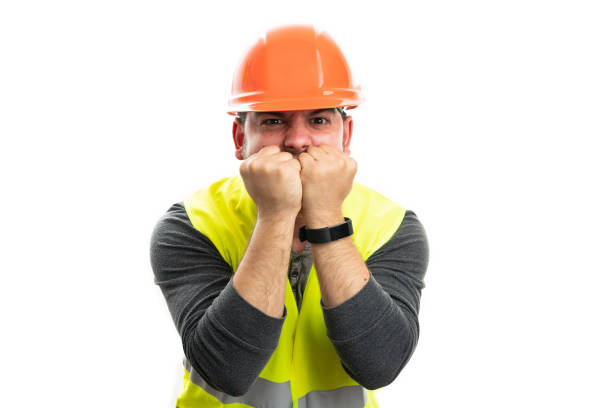 Builder man with angry desperate expression wearing fluorescent vest and orange helmet using fists to cover mouth gesture isolated on white background