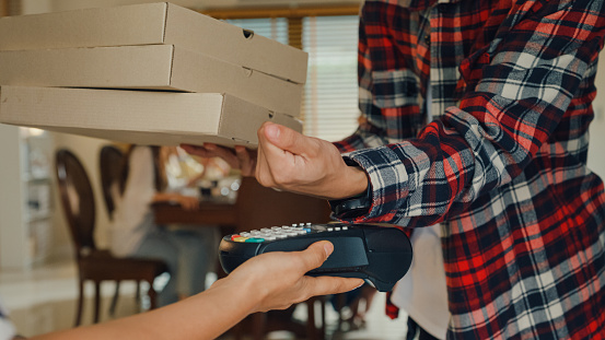 Closeup of consumer paying using smart watch and NFC technology to courier for package delivery at home. Multicultural friends having fun together college house party concept.