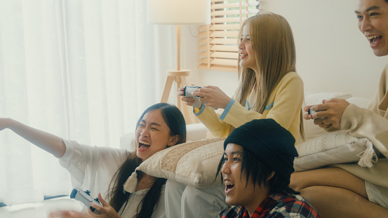 Group of young Asian people playing video games colorful party on sofa in living room at home. Multicultural friends enjoying spending together college house party concept.