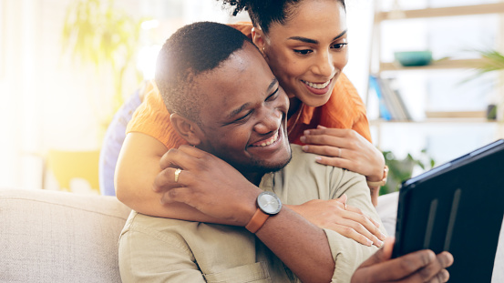 Couple, tablet and smile with hug on sofa in home living room, online browsing or web scrolling. Interracial, technology and happy black man and woman bonding on couch in lounge on social media.