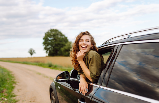 Young woman leaning out of the car window enjoying nature. Girl is resting and enjoying journey. Lifestyle, travel, tourism, active life.