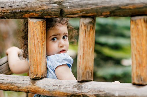 Thoughtful curly hair toddler girl sitting on handcrafted wooden bench and looking at camera sadly