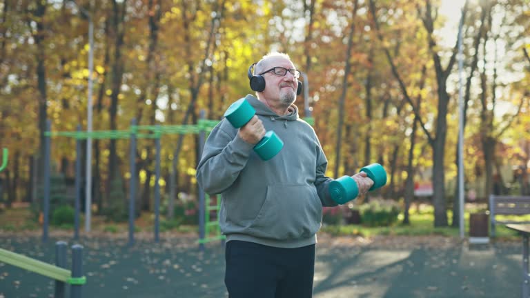 A cheerful elderly man lifts dumbbells in the park while wearing headphones and dancing to music